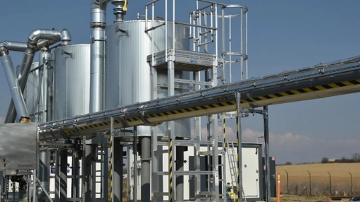 Biomethane plant with large metal tanks, overhead pipelines and an industrial conveyor beside open farmland