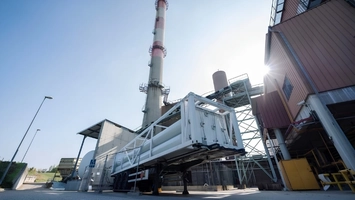 Ground-level view of a biomethane processing unit beside an industrial building with a tall chimney stack