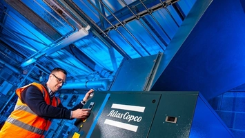 Technician in an orange high-vis vest operating an Atlas Copco compressor in a blue-lit industrial plant