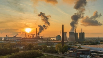 Industrial facility with large cooling towers and chimneys releasing emissions against an orange sunset sky
